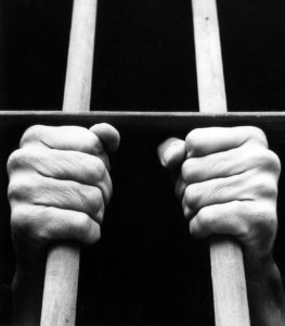 Black and white photograph showing hands of anonymous male prisoners gripping the bars to his prison cell. This is a reconstruction, use for illustrative purposes.